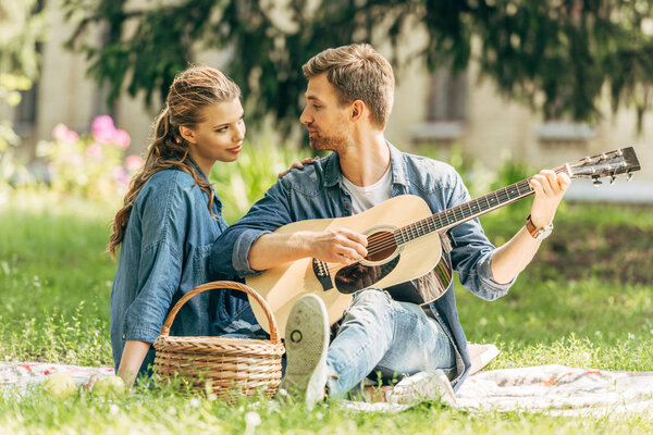handsome young man playing guitar for his smiling girlfriend during picnic at park
