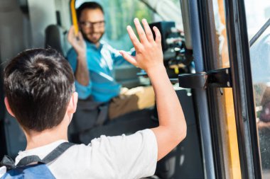 rear view of schoolboy waving to happy bus driver while entering bus clipart