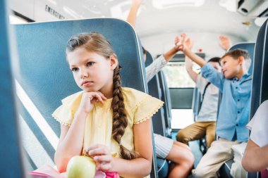 sad little schoolgirl riding on school bus while her classmates giving high five blurred on background clipart