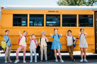 group of adorable schoolchildren standing in front of school bus and showing thumbs up clipart