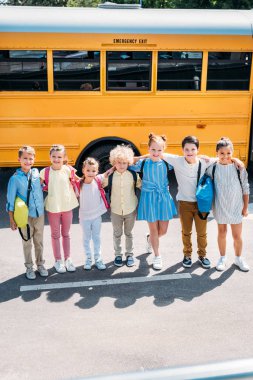 group of happy schoolchildren standing in front of school bus and looking at camera clipart