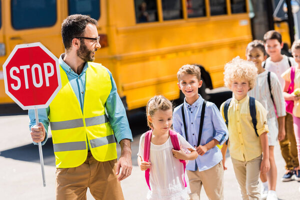 красивый дорожный охранник пересекает дорогу со школьниками перед школьным автобусом
