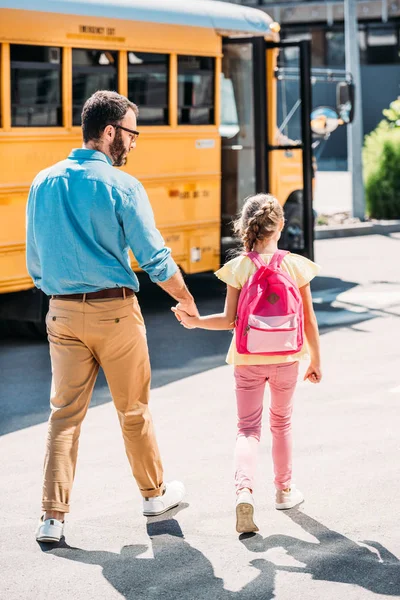 父と娘は手を繋いでいると学校のバスに歩いてバックパックの背面図 — ストック写真