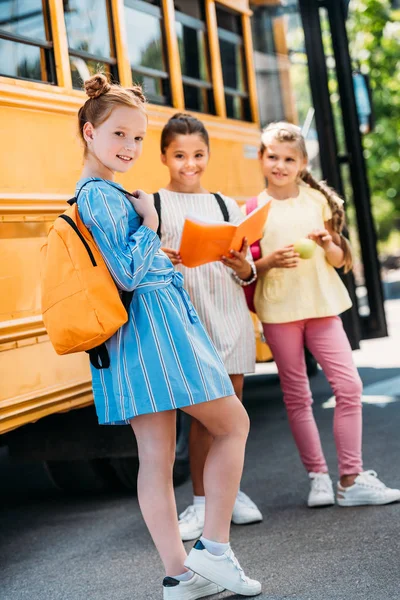 Szczęśliwy Mało Uczennice Notebook Ogląda Aparat Przed Autobus Szkolny — Darmowe zdjęcie stockowe