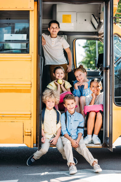 group of cute schoolchildren sitting on stairs of school bus and looking at camera