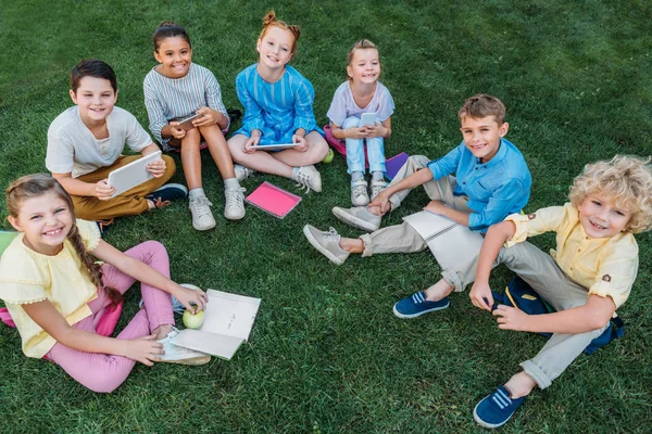 本デバイスと草の上に座って幸せな小学生のグループのハイアングル — ストック写真