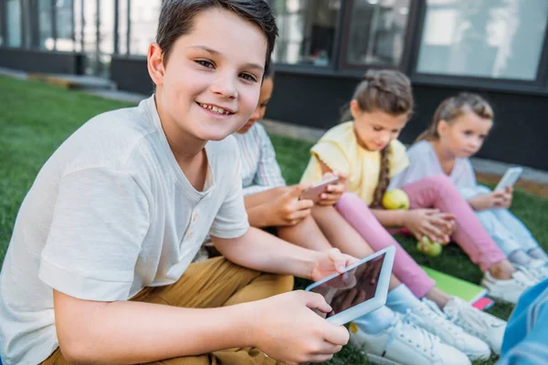 Sonriente Colegial Usando Tableta Mientras Sentado Hierba Con Compañeros Clase — Foto de Stock