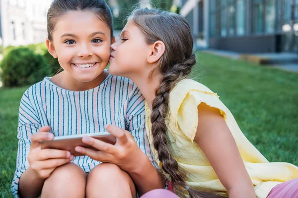 Adorable Little Uczennica Smartphone Patrząc Kamery Podczas Jej Przyjaciel Całuje — Zdjęcie stockowe