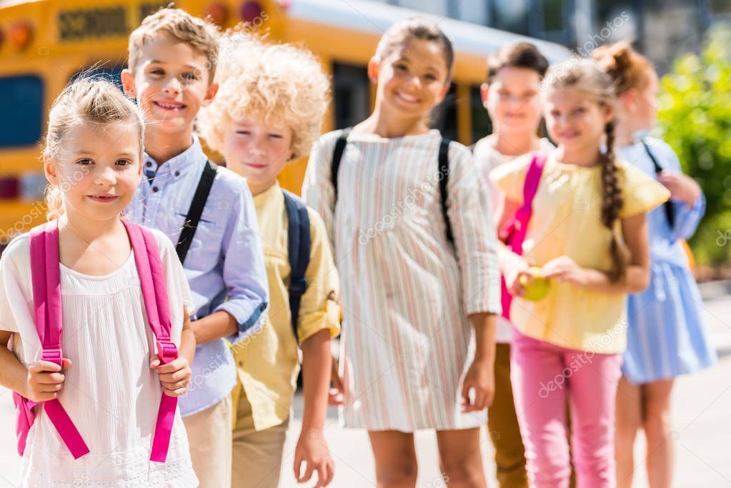 group of adorable schoolchildren standing in row in front of school bus and looking at camera