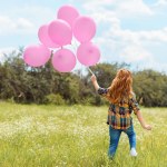 Back view of child with pink balloons standing in summer field with blue sky on background
