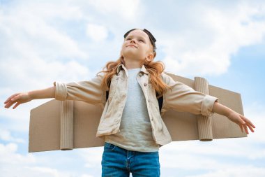 low angle view of child with paper plane wings and protective eyeglasses with outstretched arms against blue cloudy sky clipart