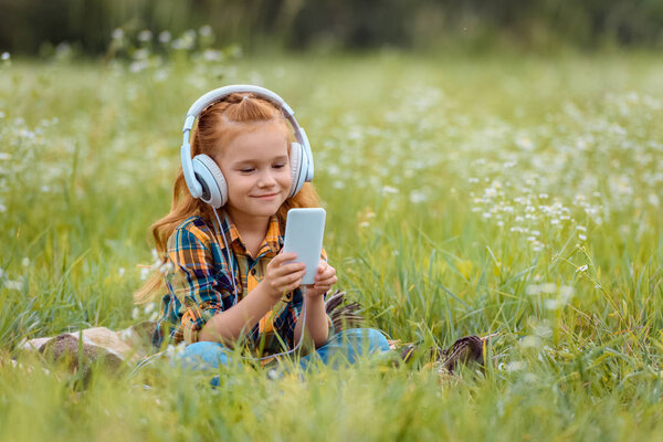улыбающийся ребенок в наушниках с помощью смартфона, отдыхая на одеяле на лугу с дикими цветами
