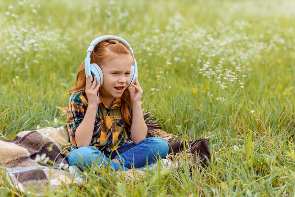 cute kid listening music in headphone while sitting on blanket in field with wild flowers