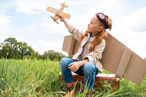 kid in pilot costume with wooden toy plane in hand sitting on retro suitcase in field