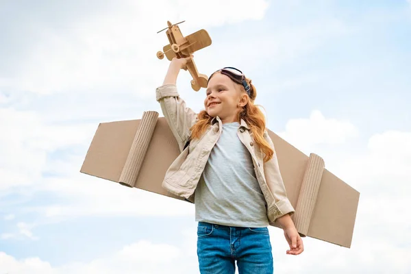 low angle view of child in pilot costume holding wooden toy plane against blue cloudy sky