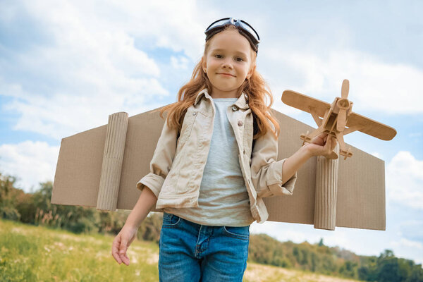portrait of smiling red hair kid in pilot costume holding wooden plane in summer field