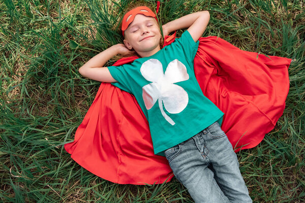 над головой вид ребенка с закрытыми глазами в красном супергеройском плаще и маске, лежащих на зеленой траве
