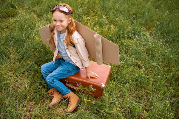 High angle view of smiling child with paper plane wings and protective eyeglasses resting on retro suitcase in meadow — Stock Image High Angle View Smiling Child Paper Plane Wings Protective Eyeglasses Royalty Free Stock Photos