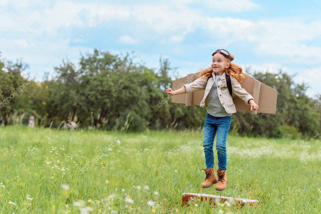 smiling kid in pilot costume jumping from retro suitcase in summer field
