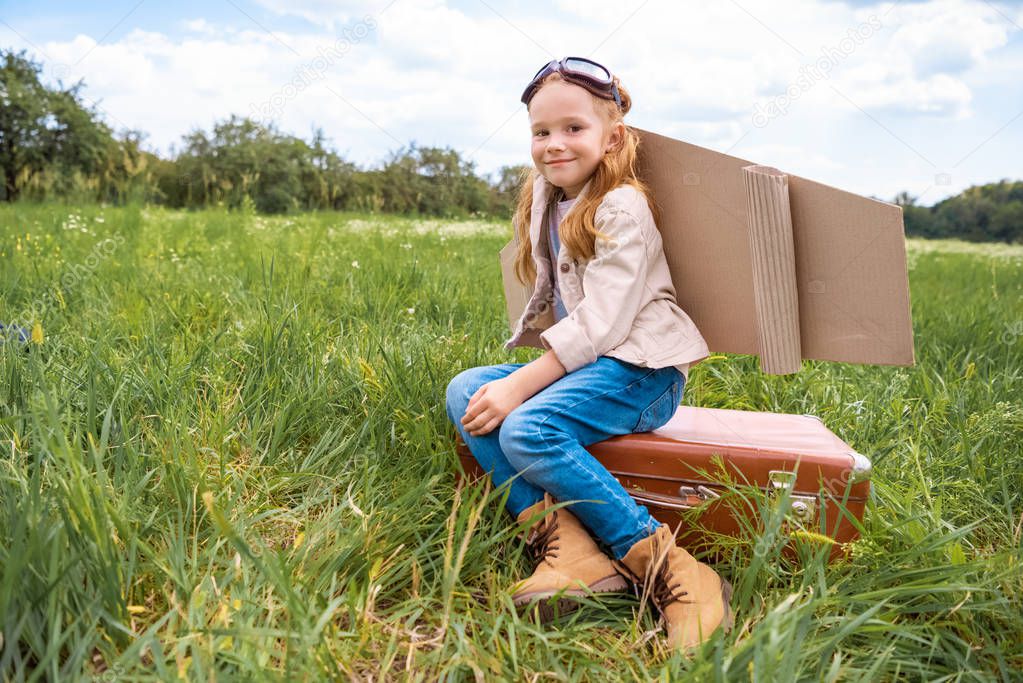 smiling cute kid in pilot costume sitting on retro suitcase in summer field
