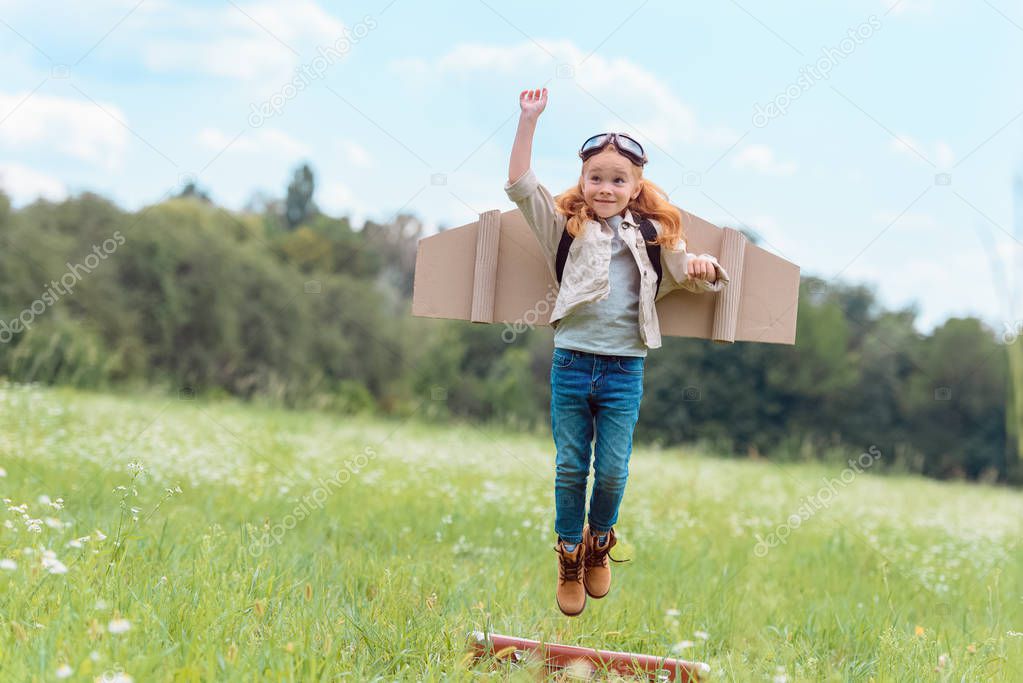 kid in pilot costume with outstretched arm jumping from retro suitcase in meadow 