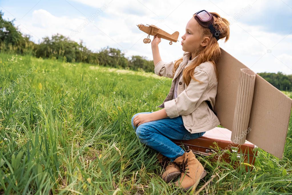 kid in pilot costume with wooden toy plane in hand sitting on retro suitcase in field