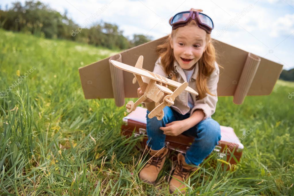 emotional kid in pilot costume with wooden toy plane in hand sitting on retro suitcase in field