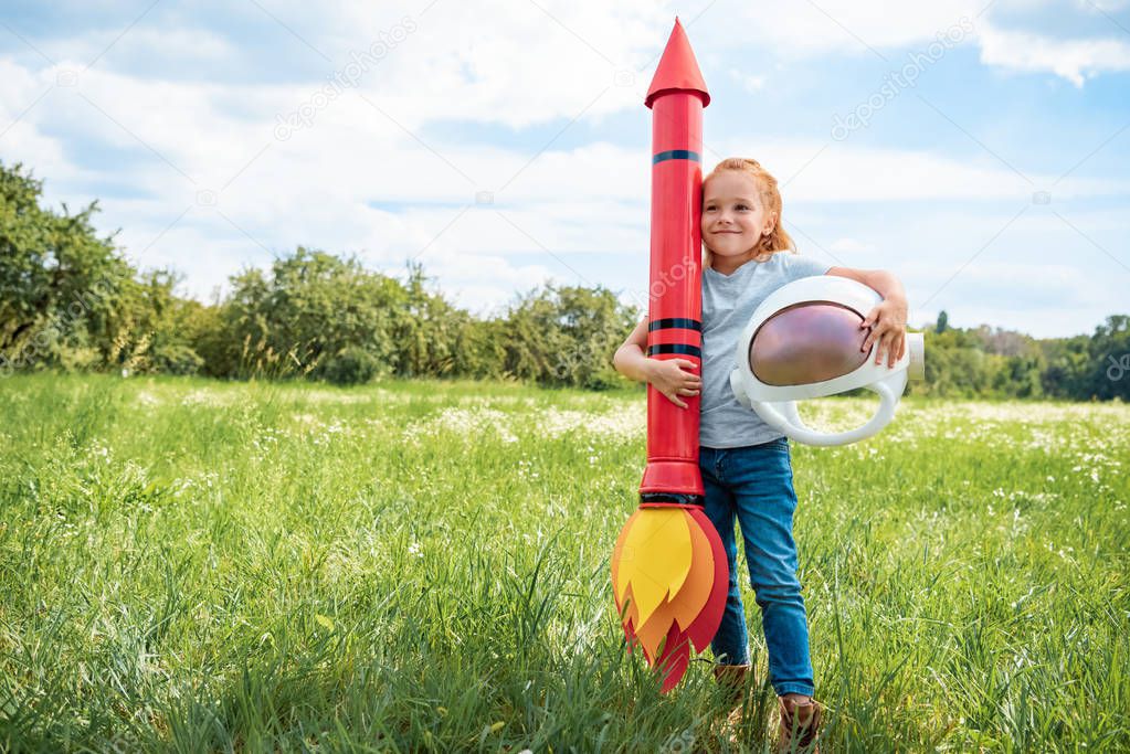 smiling red hair kid with rocket and astronaut helmet standing in summer field