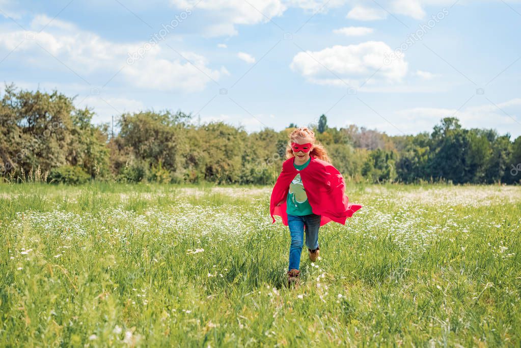 little kid in red superhero cape and mask running in meadow on summer day