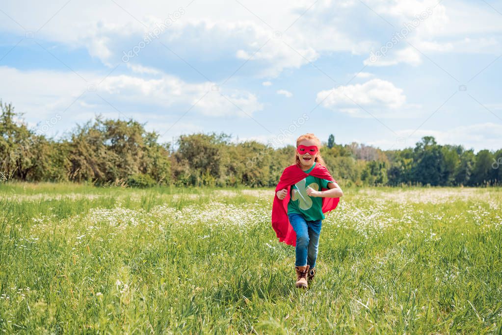 happy kid in red superhero cape and mask running in meadow on summer day