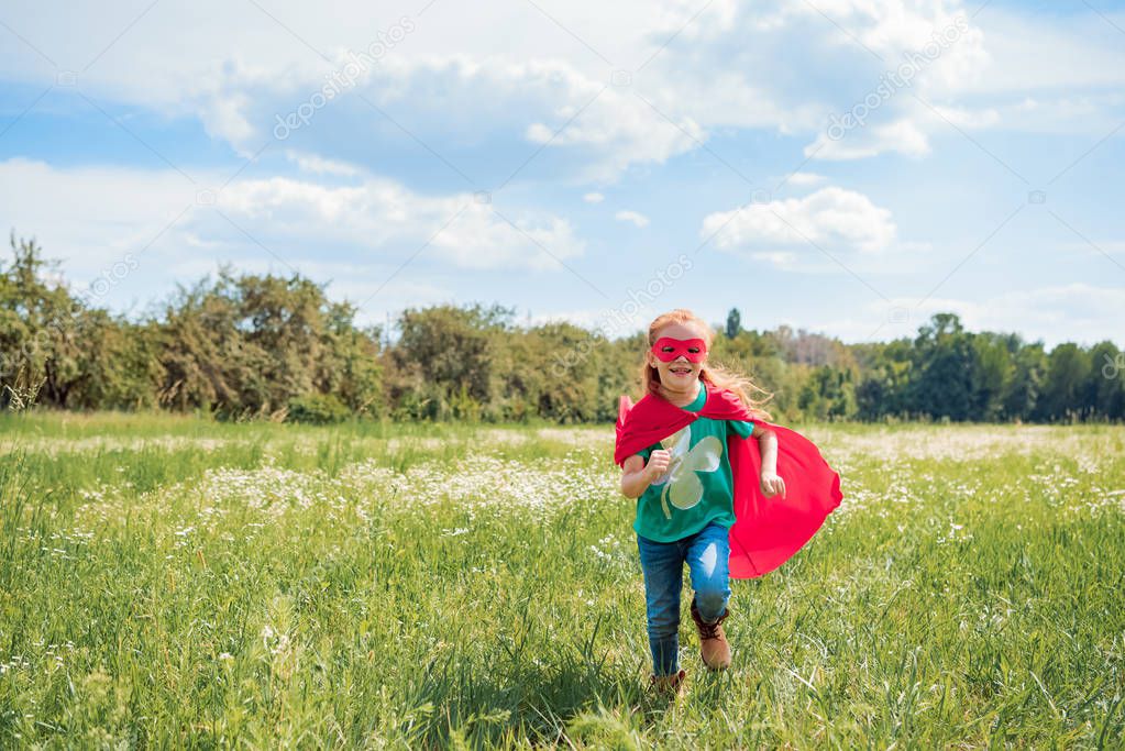 cheerful kid in red superhero cape and mask running in meadow on summer day