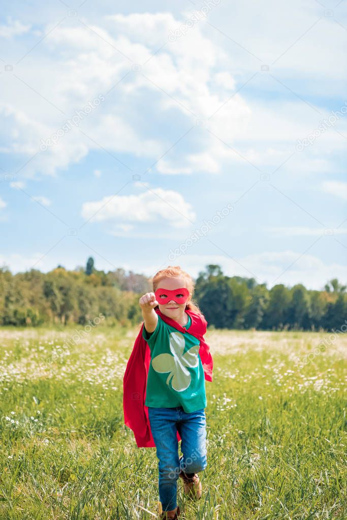 little kid in red superhero cape and mask with outstretched arm running in meadow on summer day