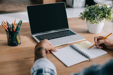 kalemler ve boş ekran ile dizüstü bilgisayar ile defter işyerinde yazma adamın kısmi görünümü