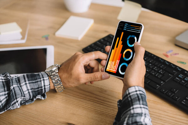 cropped shot of seo using smartphone with statistics charts on screen at workplace