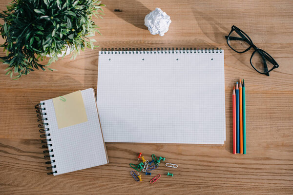 top view of notebooks, paper clips, pencils, eyeglasses and plant on wooden tabletop in office