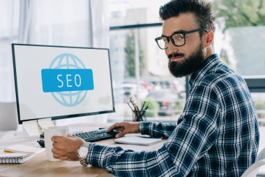 bilgisayar ve kameraya bakarak ile işyerinde oturan genç başarılı seo Yöneticisi