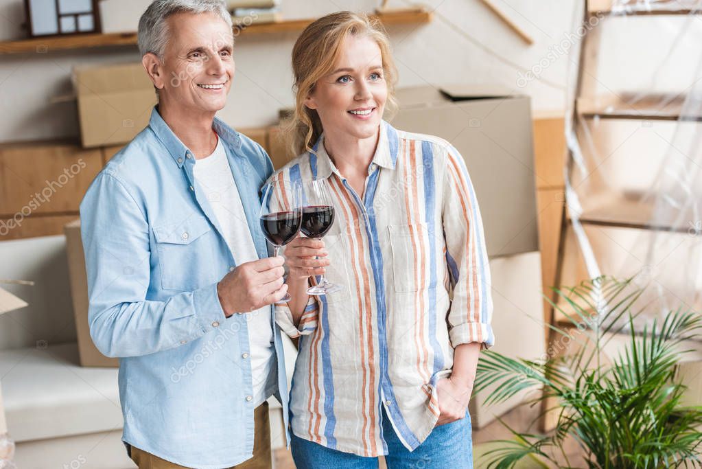 happy senior couple holding glasses of wine and looking away in new house