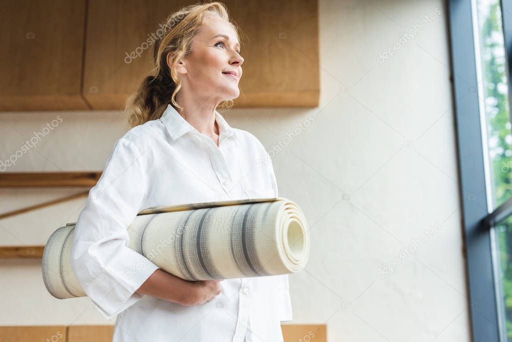 low angle view of beautiful smiling mature woman holding yoga mat and looking away