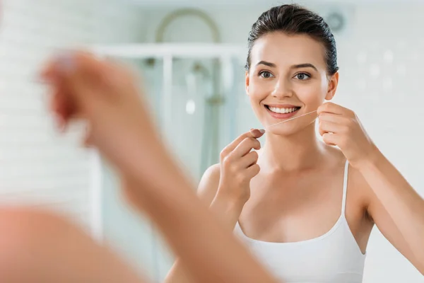 微笑的年轻妇女拿着牙线并且看镜子在洗手间 — 图库照片