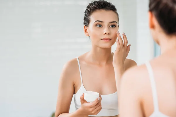 Hermosa Chica Aplicando Crema Facial Mirando Espejo Baño — Foto de Stock