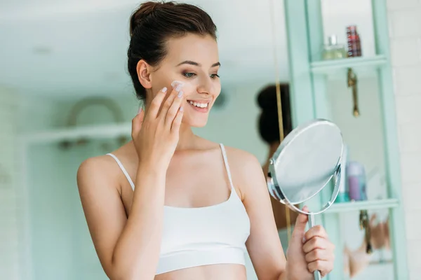 Bela Menina Sorridente Segurando Espelho Aplicando Creme Facial Banheiro — Fotografia de Stock