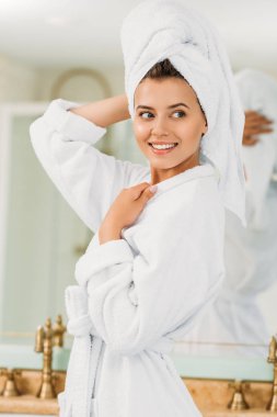 beautiful smiling girl in bathrobe and towel on head looking away in bathroom clipart