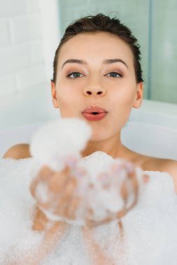 beautiful young woman blowing foam and looking at camera in bathroom clipart