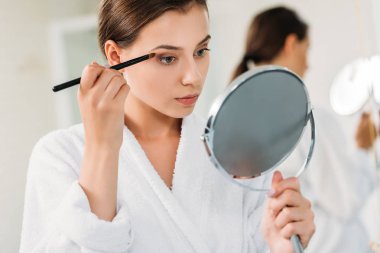 beautiful young woman holding mirror and applying eyeshadow  clipart