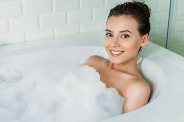высокий угол зрения красивой счастливой девушки расслабляясь в ванне с пеной и улыбаясь на камеру
