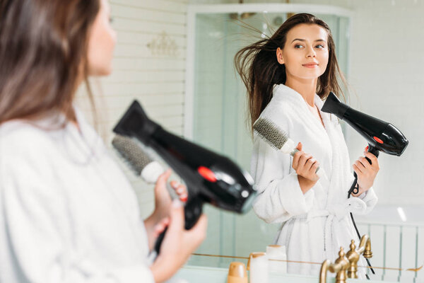 beautiful young woman holding hair brush and drying hair at mirror in bathroom