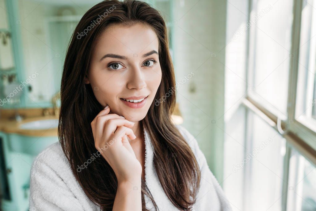 beautiful brunette girl in bathrobe smiling at camera in bathroom
