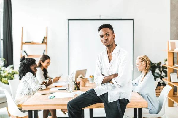 腕を組んでの背後に働く多文化の同僚とテーブルに座って物思いにふけるアフリカ系アメリカ人実業家の肖像画 — ストック写真