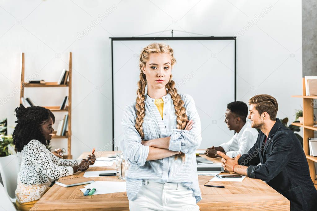 portrait of upset businesswoman with arms crossed looking at camera and colleagues working behind in office