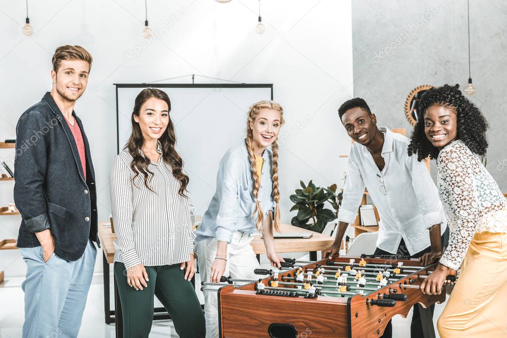 multicultural smiling business people looking at camera while playing table football in office